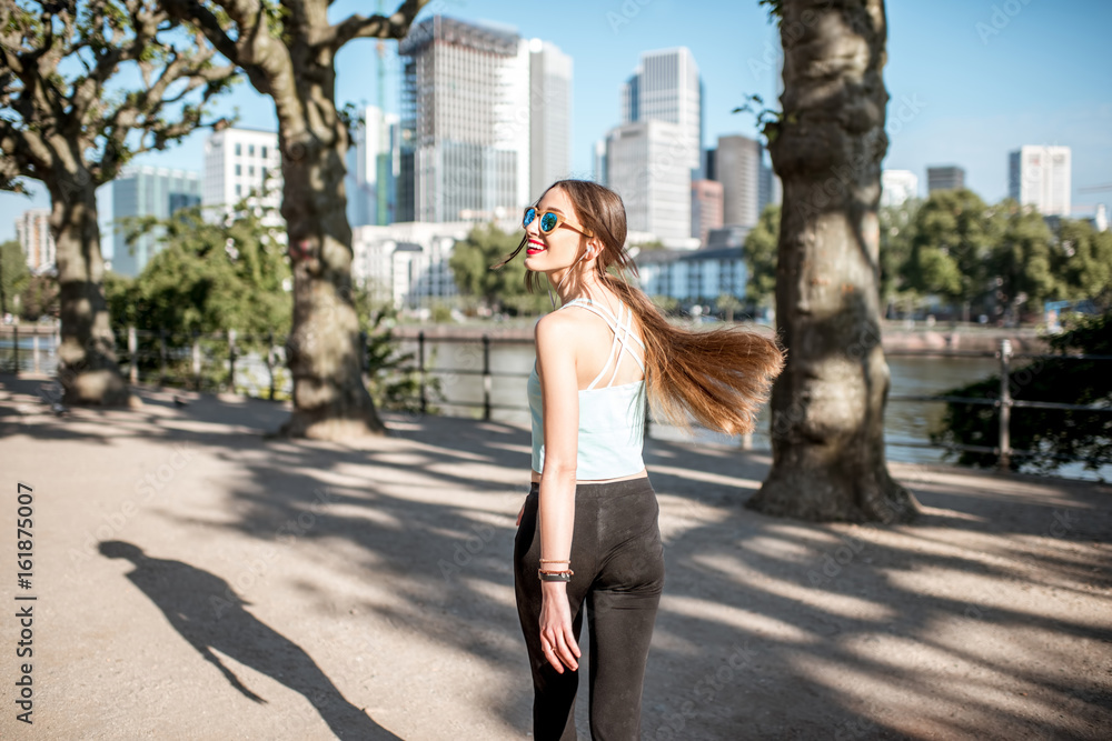 Young woman in sportswear walking and looking back in the park with skyscrapers on the background in