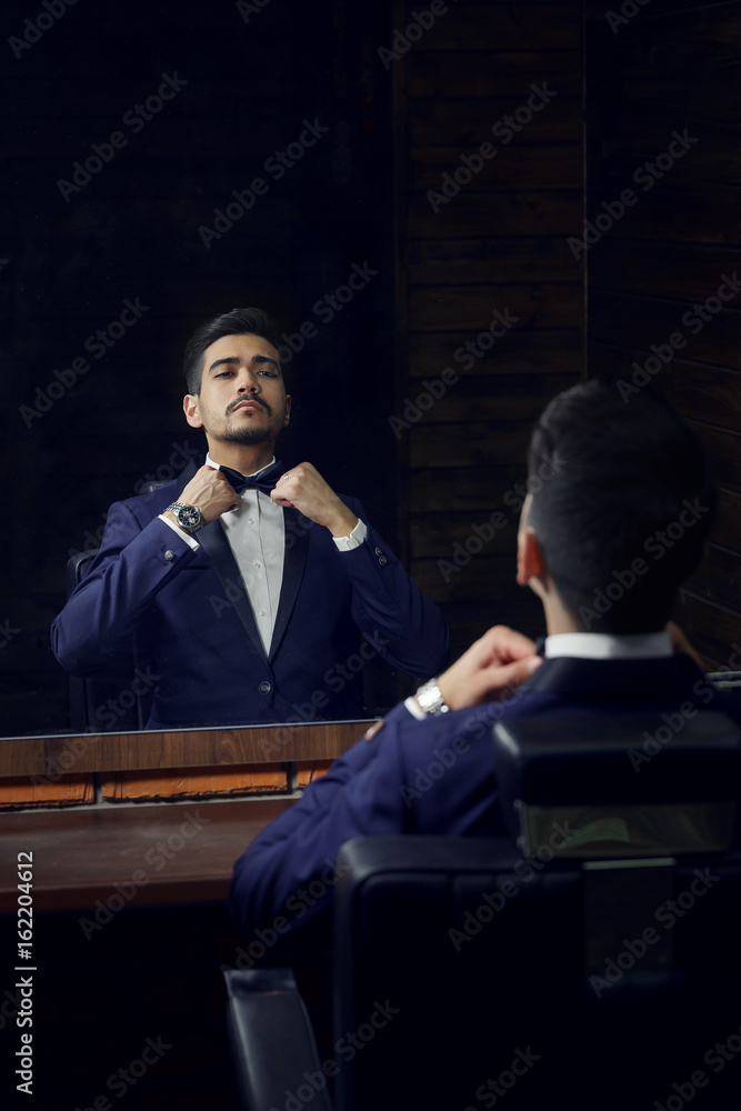 Confident man in blue suit and butterfly sitting in chair looking in the mirror in barbershop