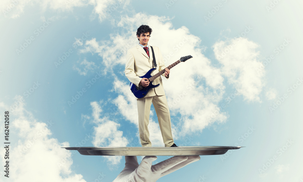 Businessman on metal tray playing electric guitar against blue sky background