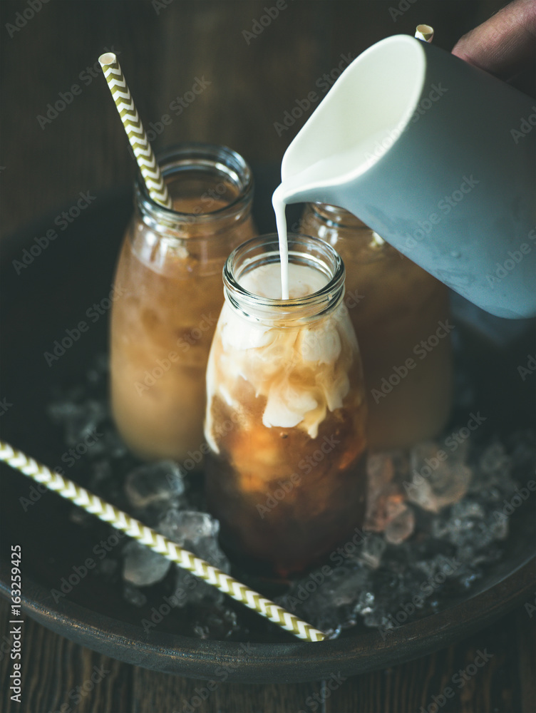 Refreshing summer drink. Cold Thai iced tea in glass bottles with milk pouring from jug on plate ove