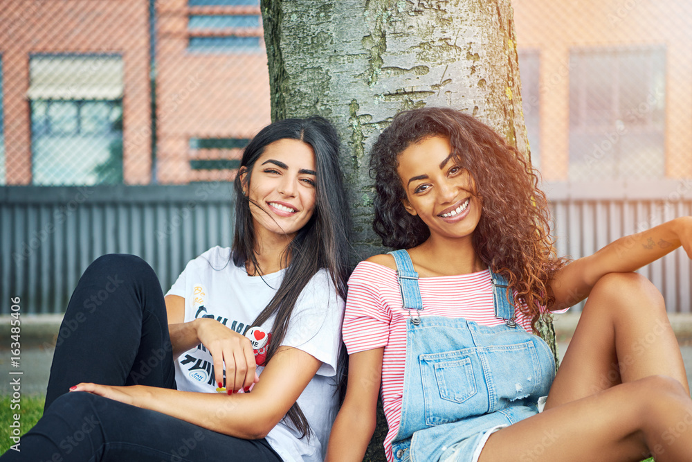 Two smiling young friends sitting under a tree together