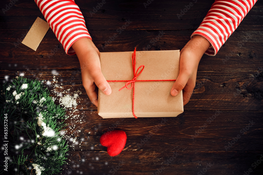 A woman hands holding a Christmas gift with a red ribbon and snow over on a wooden table. Handmade C