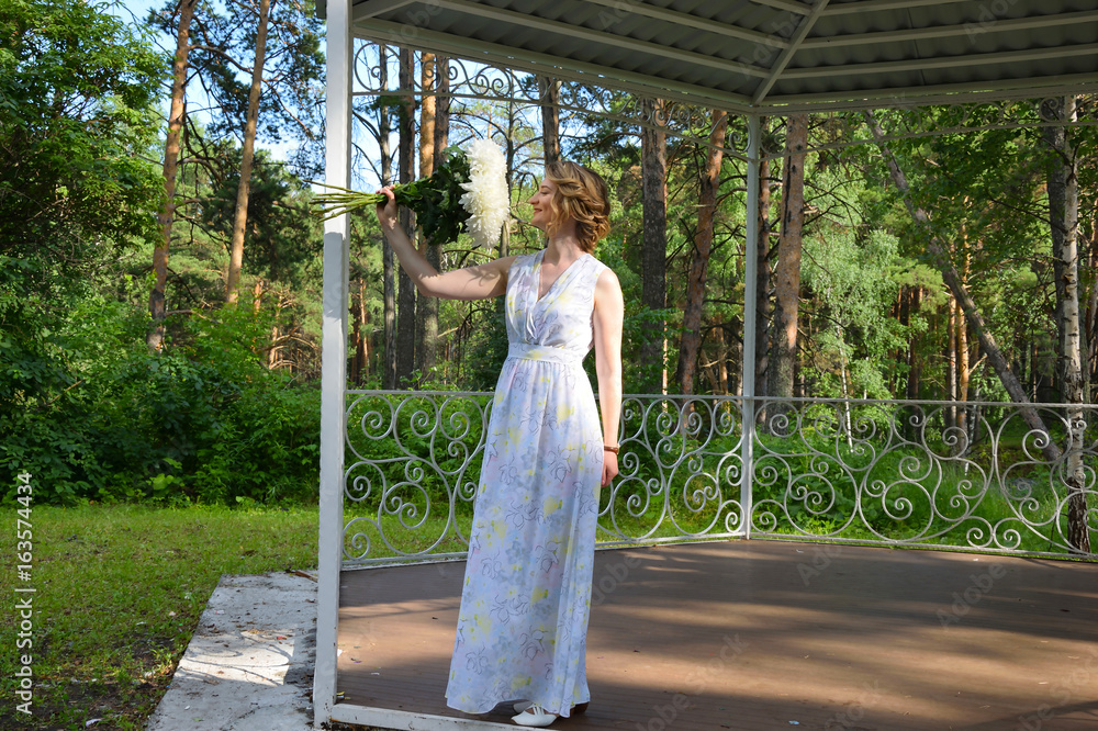 A happy girl in a long dress with flowers is standing in a forest arbor.