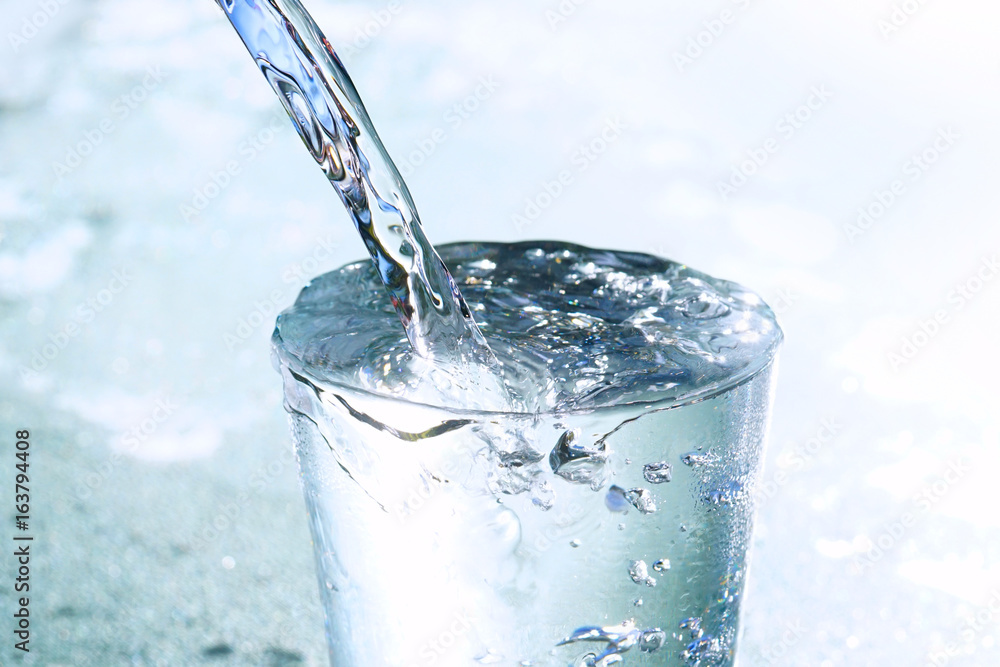 A stream of clean cold transparent water pours into a glass beaker close-up of a macro on a light ba