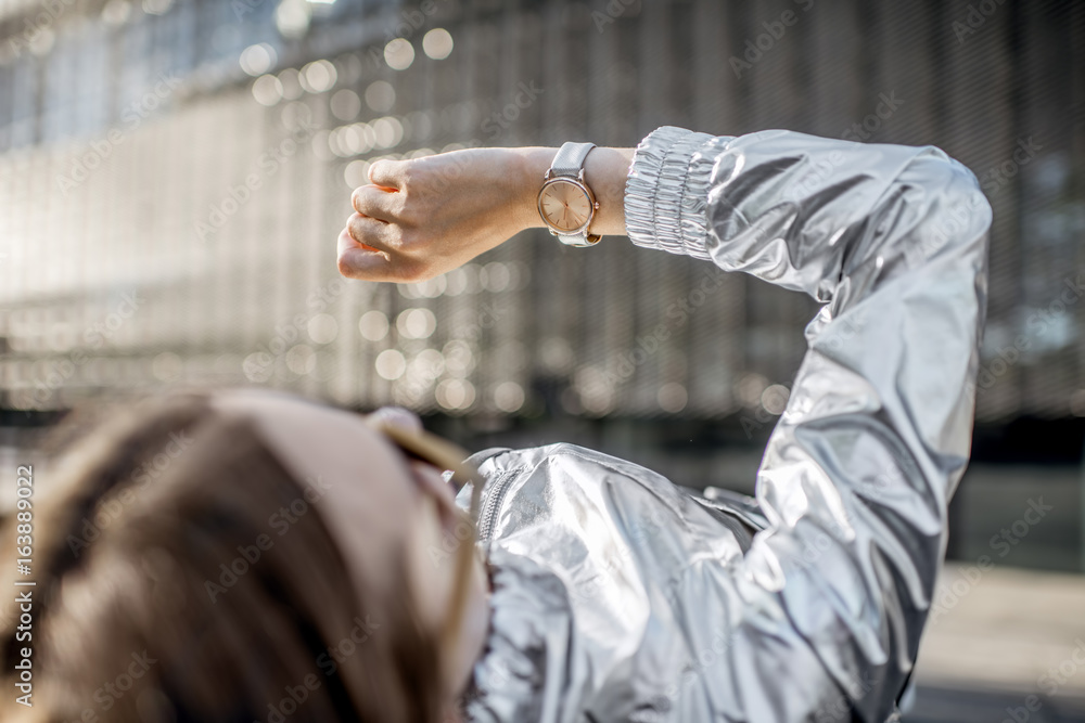 Stylish woman in silver jacket watching the clock lying outdoors at the modern environment