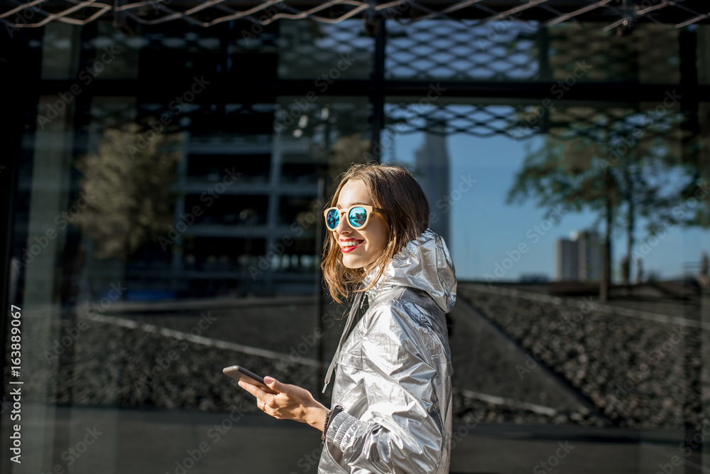 Lifestyle portrait of a stylish woman in silver jacket walking with phone on the modern facade backg