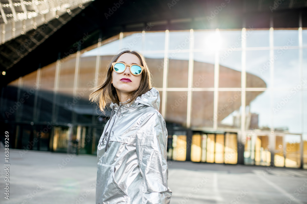 Lifestyle portrait of a stylish woman in silver jacket walking outdoors on the modern architectural 