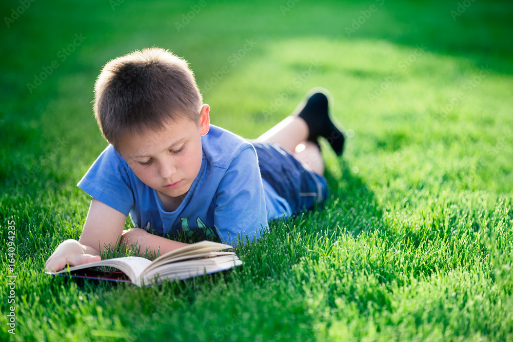 Boy reading book, lying down on green grass