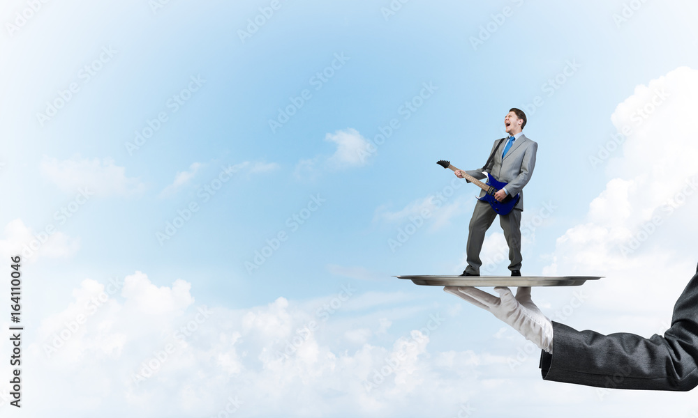 Businessman on metal tray playing electric guitar against blue sky background