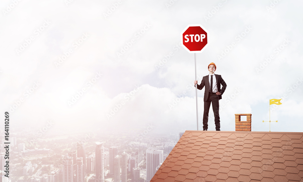 Caucasian businessman on brick house roof showing stop road sign. Mixed media