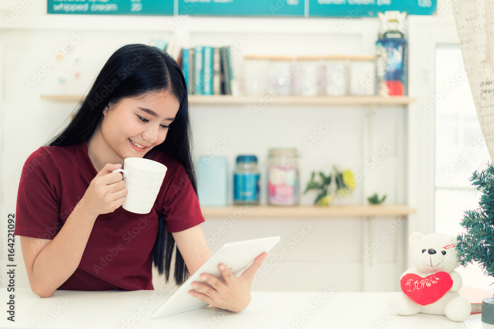 年轻的亚洲微笑女人在咖啡馆喝咖啡，手里拿着一台平板电脑。