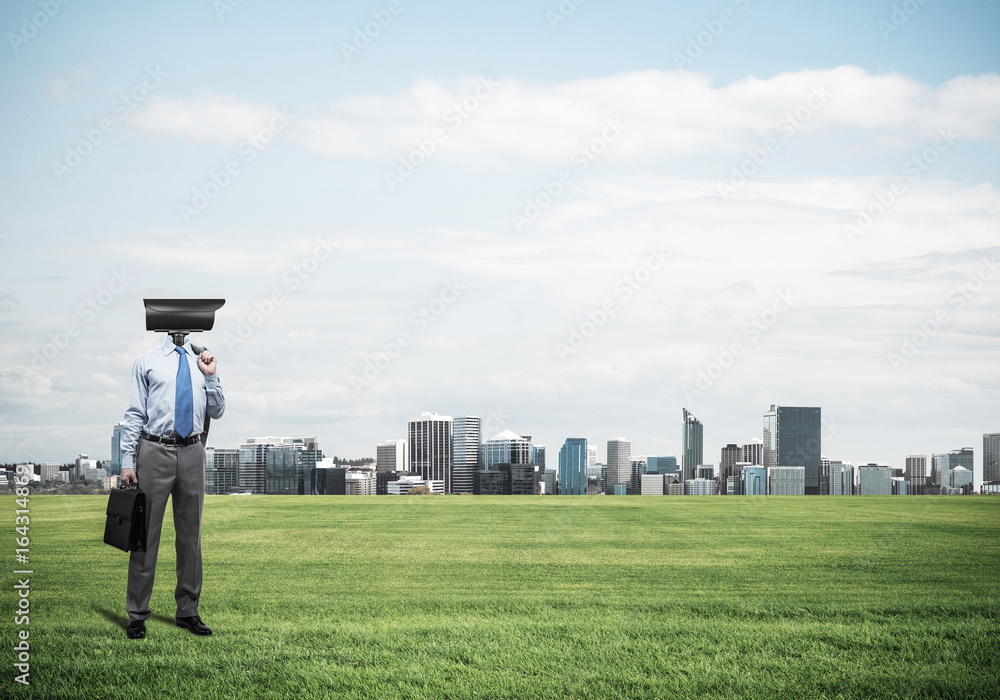 Camera headed man standing on green grass against modern citysca