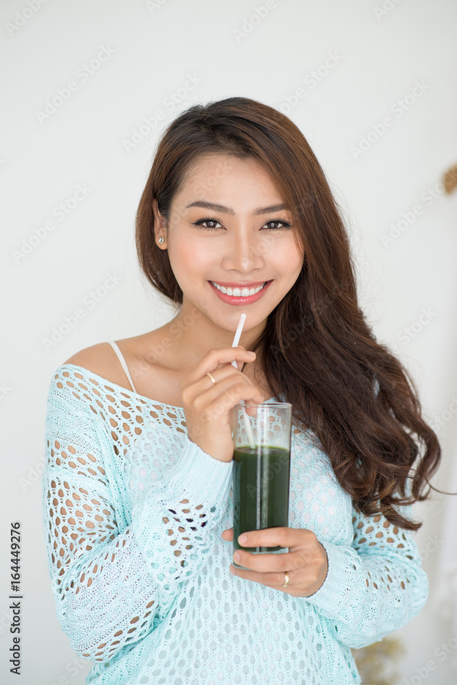 Pretty young asian woman drinking green fresh vegetable juice or smoothie from glass at home