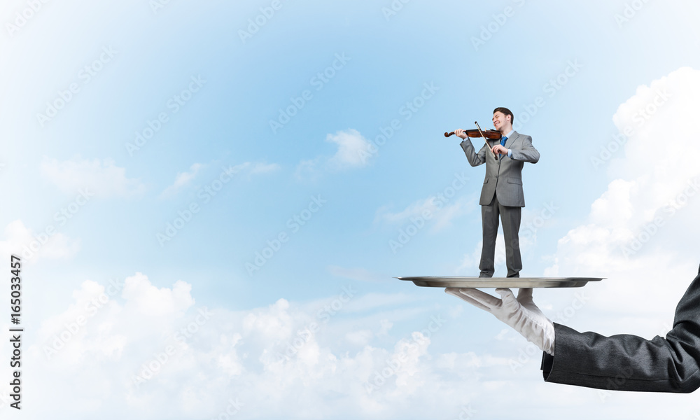 Businessman on metal tray playing violin against blue sky background