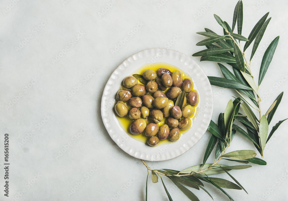 Pickled green Mediterranean olives in virgin olive oil on white ceramic plate and green olive tree b