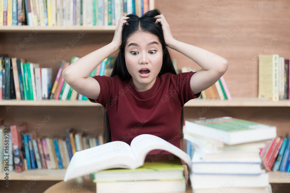 Young Asian student under mental pressure while reading book preparing examination in library at uni