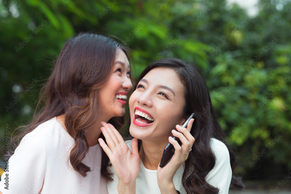 Young asian woman whispering secret into a friends ear while shes on call