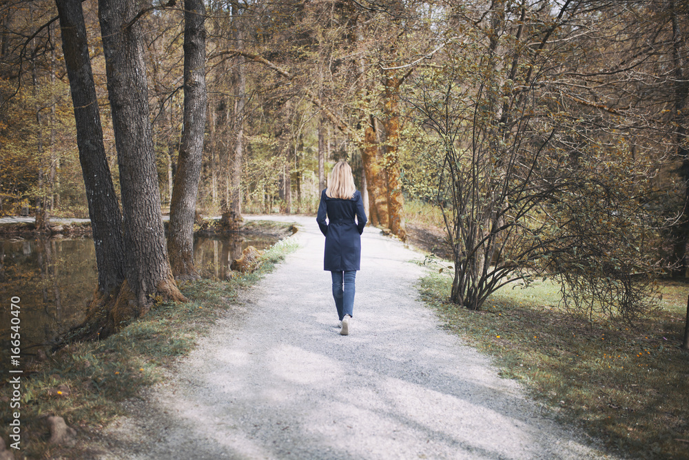 Blonde woman in blue elegant coat walks alone in the sunny seasonal park with lake and trees.