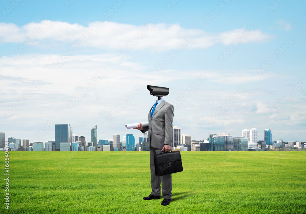 Camera headed man standing on green grass against modern cityscape