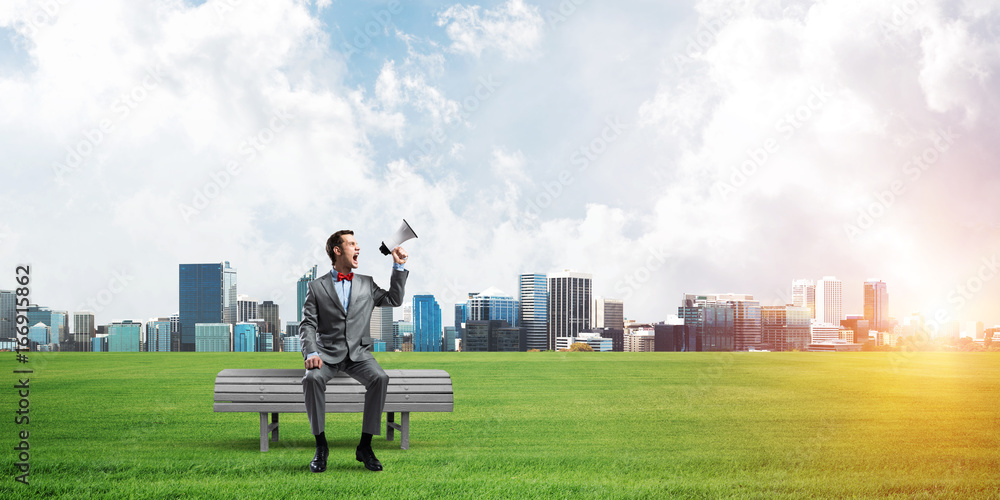 Businessman in summer park announcing something in loudspeaker and cityscape at background