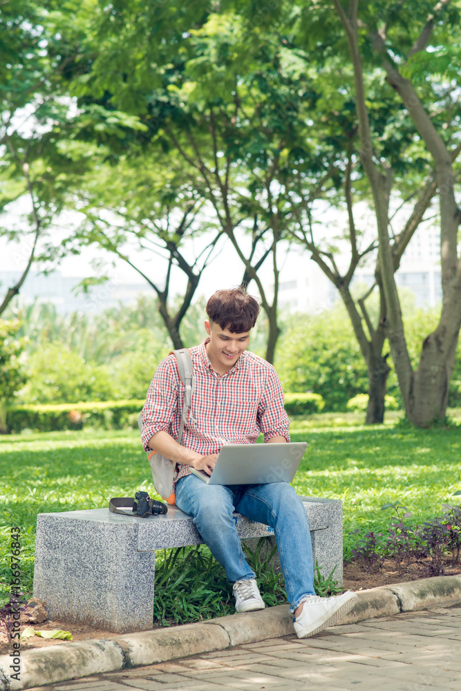 Young man sitting on bench in park, reading book, smiling and looking into the camera