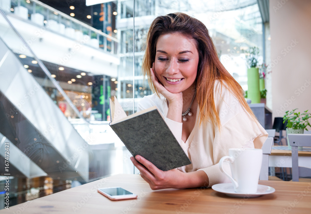 Relaxed smiling girl reading book in cafe at shopping mall