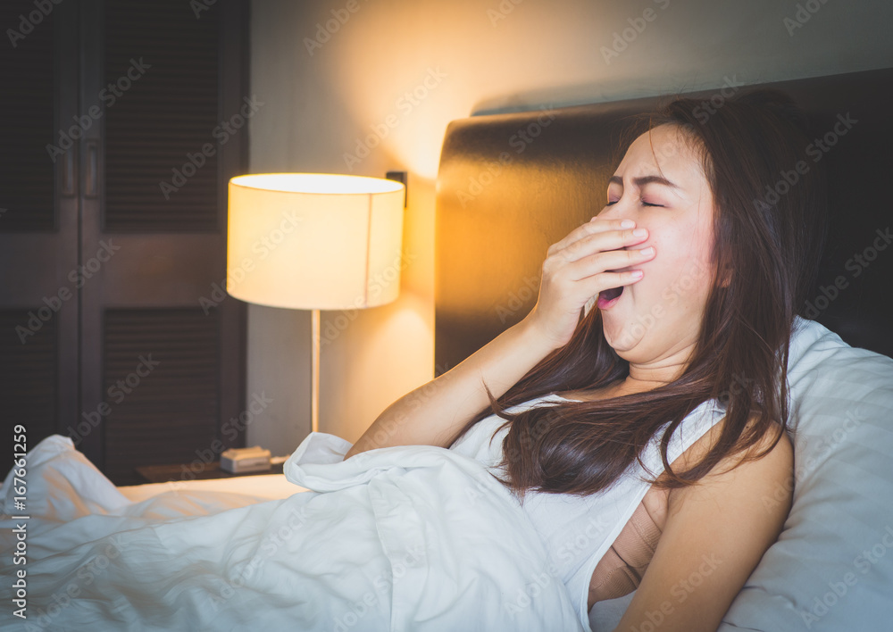 年轻漂亮的亚洲女人在卧室的白色床上打哈欠，感到昏昏欲睡。