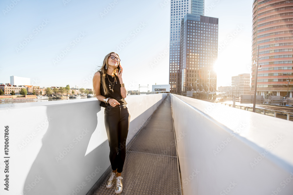 Lifestyle portrait of a stylish woman standing with phone on the modern bridge with skyscrapers on t