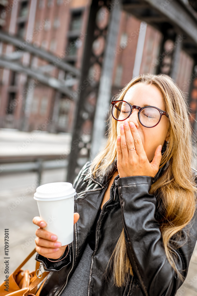 一位穿着皮夹克、拿着咖啡杯在户外打哈欠的疲惫女商人的生活方式肖像