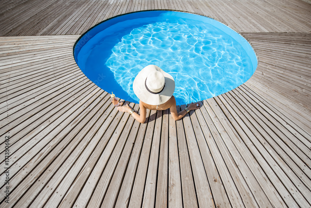 Woman in big sunhat with cocktail drink relaxing at the round swimming pool with blue water outdoors