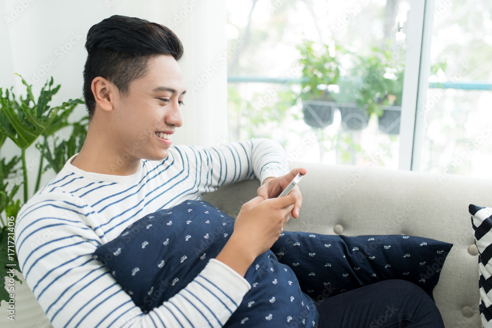 Young handsome asian man using modern mobile smart phone sitting on a couch at home