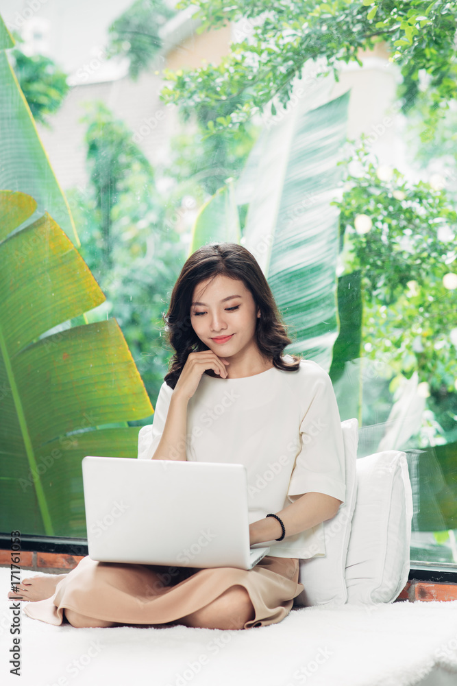 Beautiful young smiling woman working on laptop while sitting on bed at home near big window.