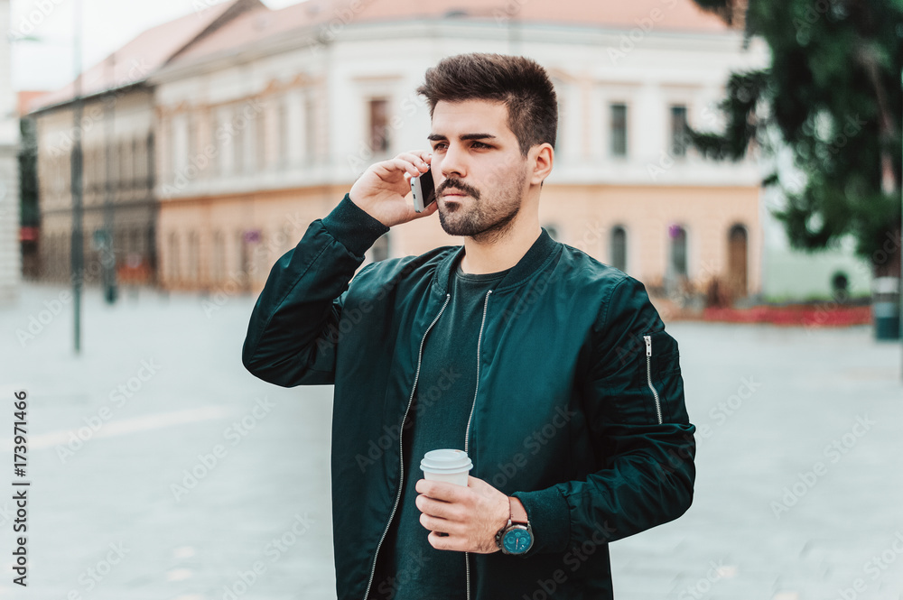 Stylish young bearded man in casual outfit is talking on his phone outside.