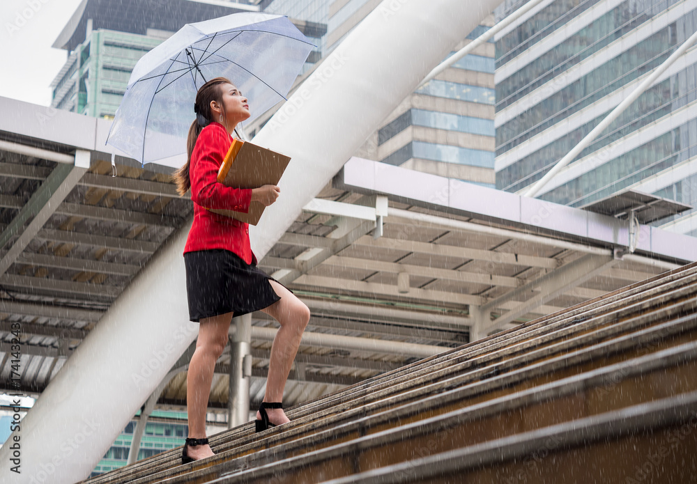 年轻的亚洲女商人拿着商业文件和雨伞在城市里雨中行走