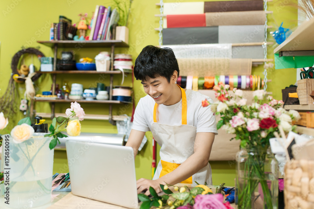 快乐的亚洲男花商在花店柜台使用笔记本电脑