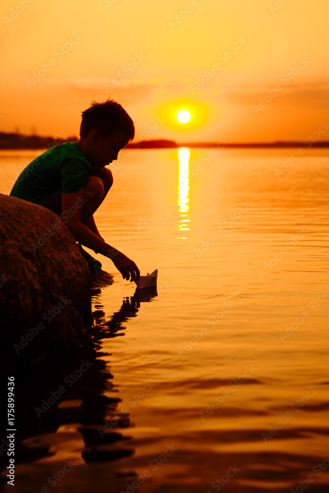 Little boy launch paper ship on the water. Beautiful summer sunset. Paper boat. Origami.