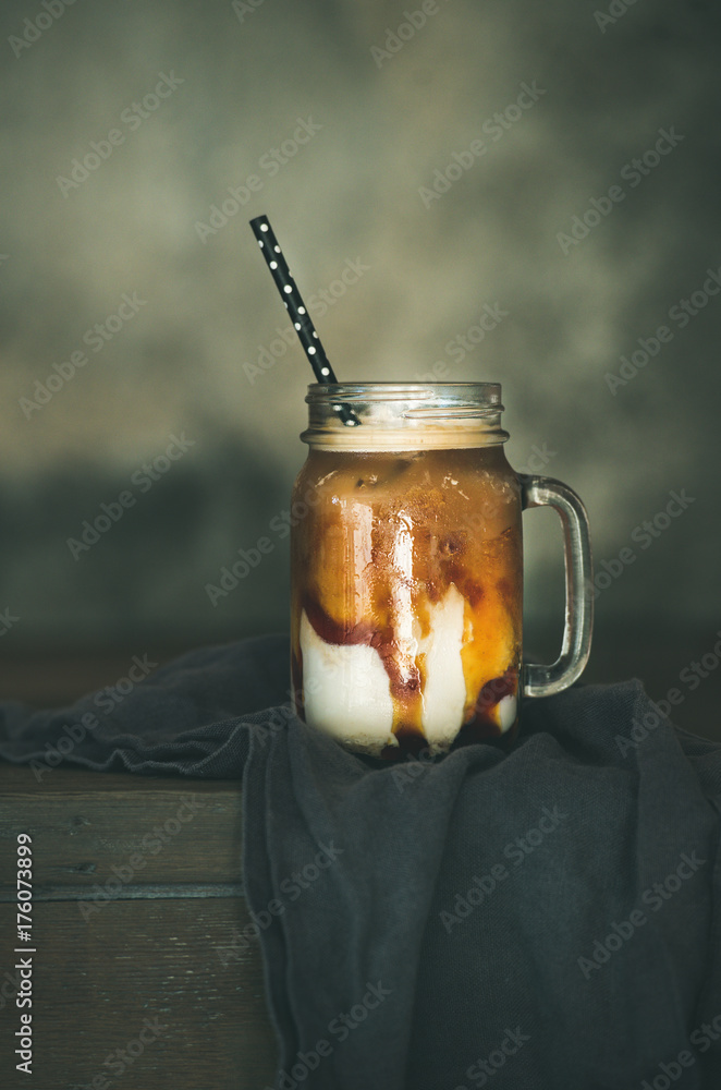 Iced caramel macciato coffee with milk in glass jar with straw on dark rustic wooden table, selectiv