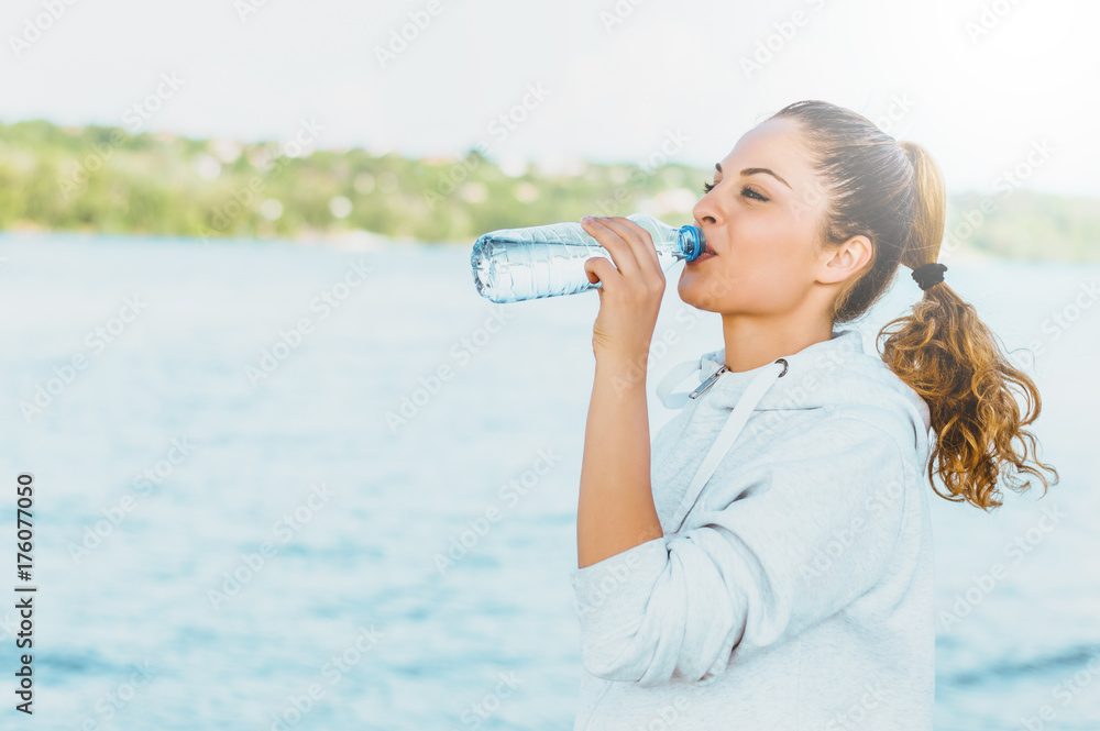 穿着运动服的漂亮女孩在海滩锻炼后喝水的侧视图。