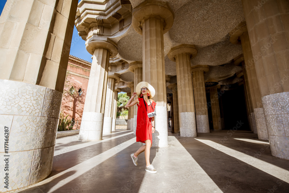 Happy woman tourist in red dress with hat walking near the columns visiting famous Guell park in Bar