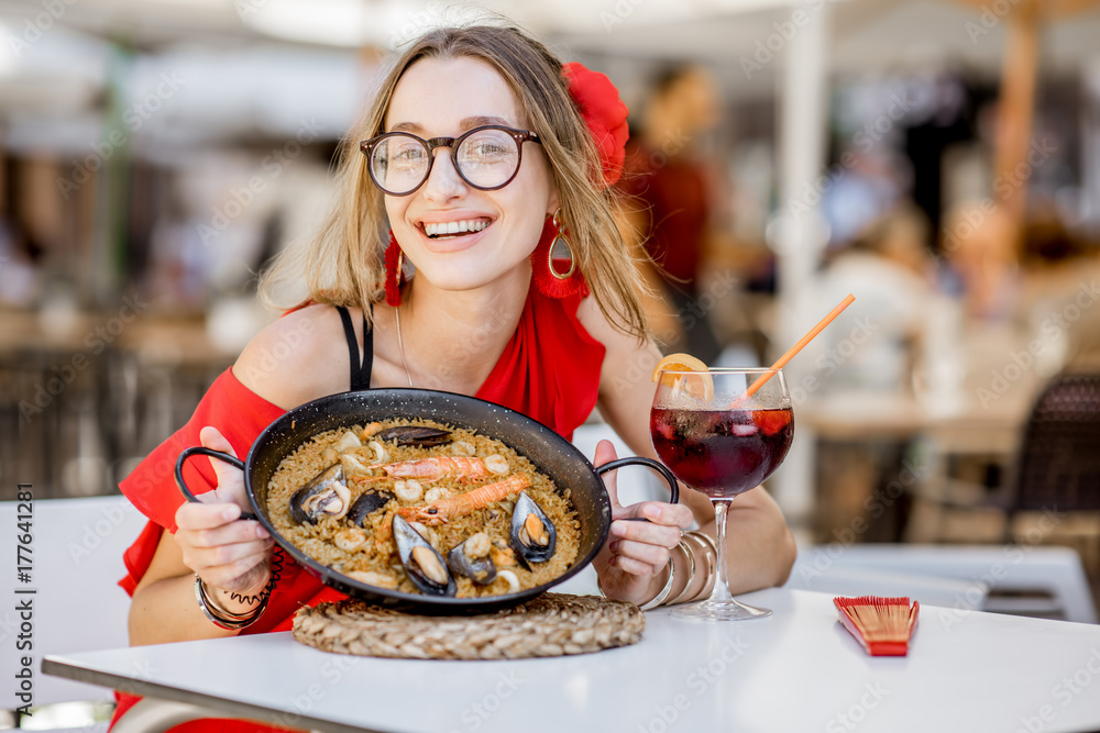 Young woman in red dress eating sea Paella, traditional Valencian rice dish, sitting outdoors at the