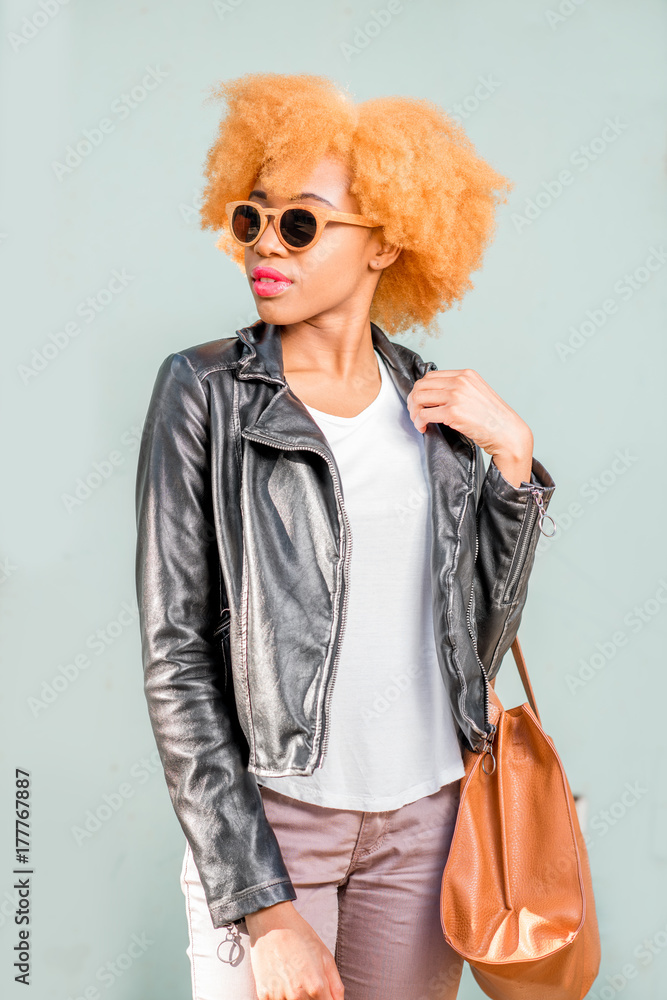 Lifestyle portrait of an african woman in leather jacket and sunglasses standing with bag on the gre