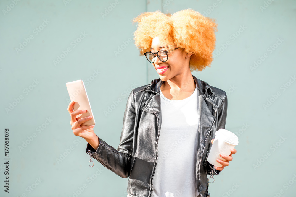 Lifestyle portrait of an african woman in leather jacket with phone and coffee cup on the green wall