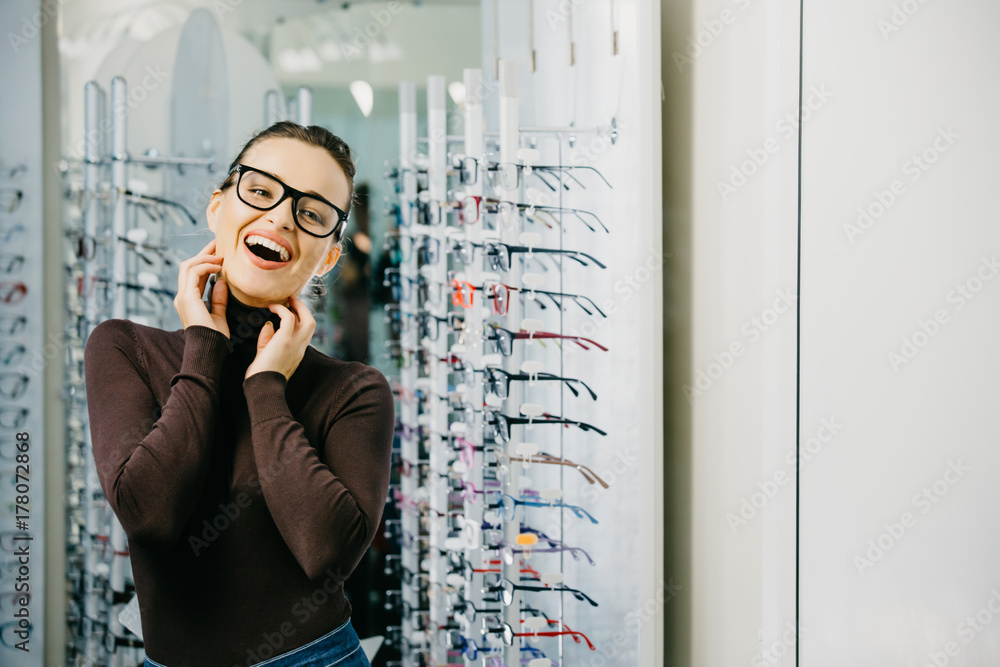 Optics. Close-up of a young beautiful girl wearing glasses, smiling near the stand. Оptical store.