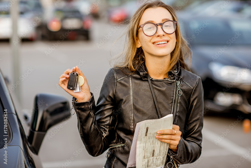 Portrait of a young happy woman with keys and rental contract standing near the car on the outdoor p