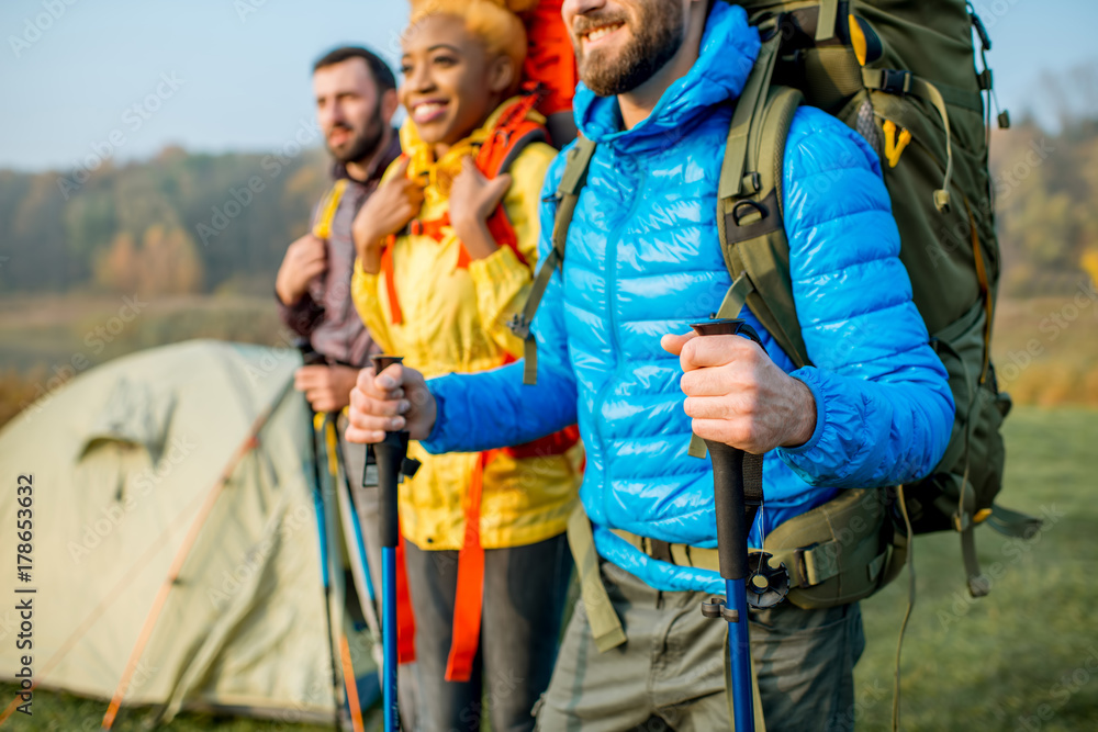 Multi ethnic friends in colorful jackets hiking with backpacks, standing near the camping on the gre