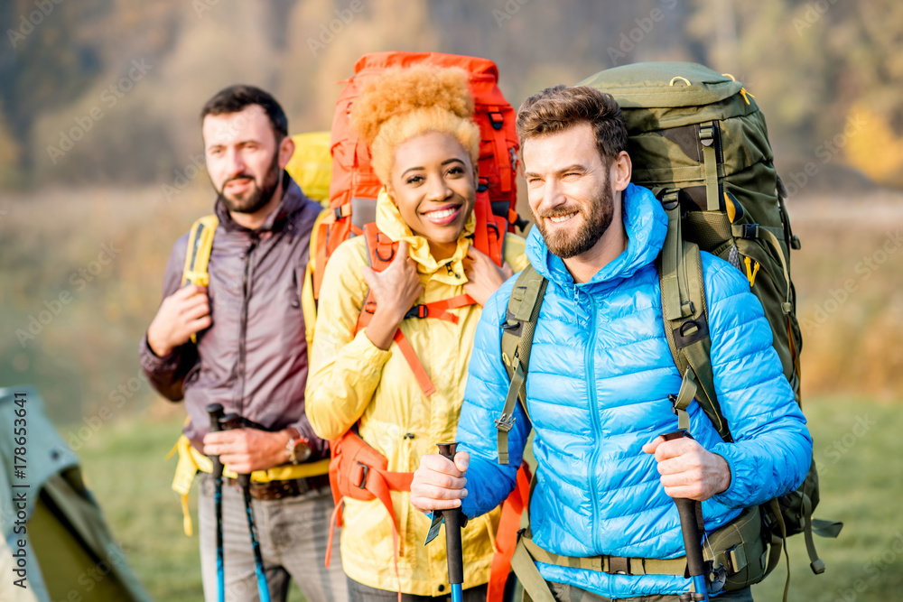 Portrait of multi ethnic friends in colorful jackets hiking with backpacks, standing near the campin