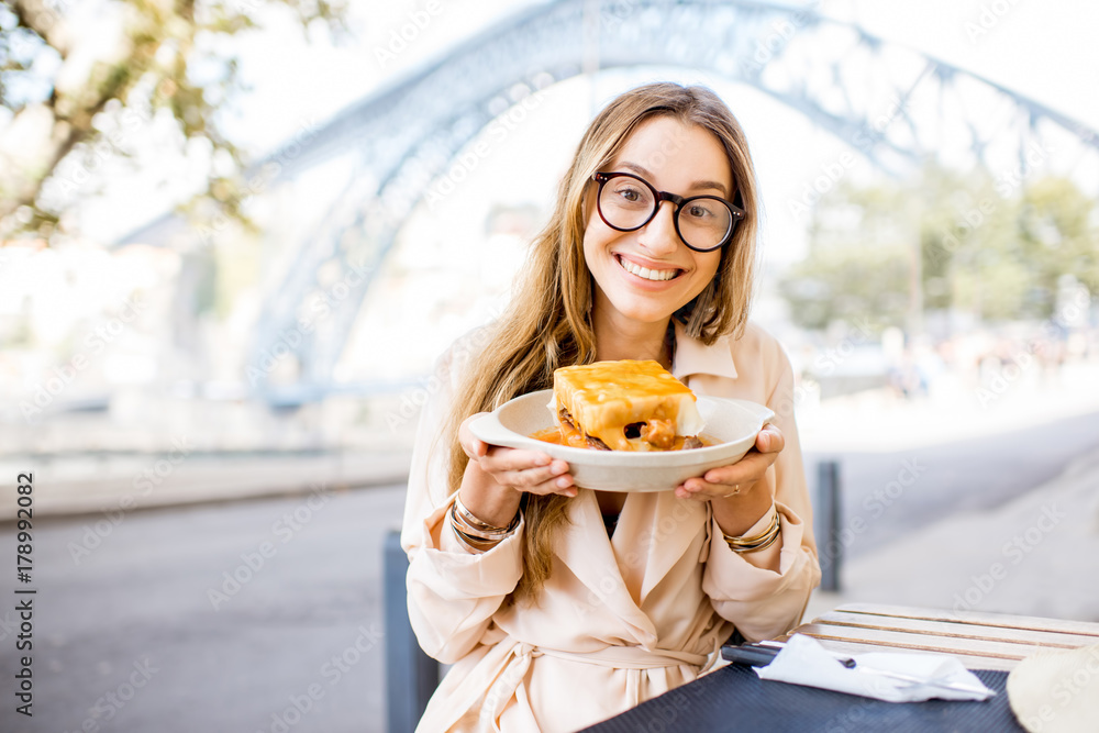 Young woman enjoying traditional portuguese meat sandwich called Francesinha, sitting at the bar wit