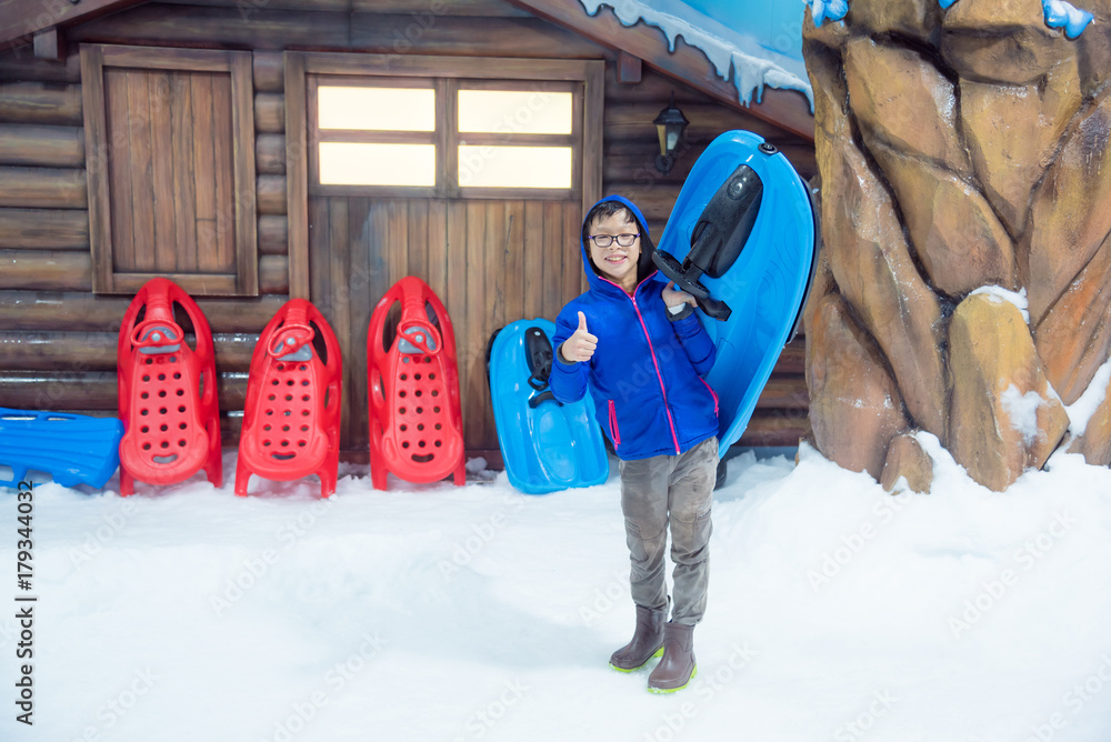 年轻的亚洲男孩拿着雪橇站在室内雪地的冰上