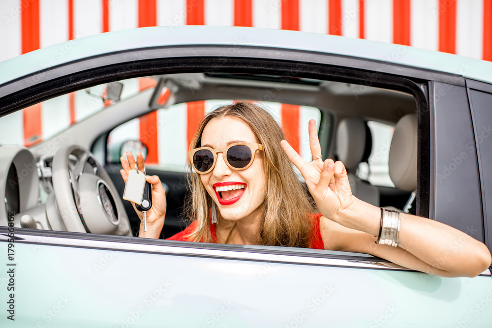 Colorful portrait of a young happy woman holding a keys pulling out the car window on the red wall b