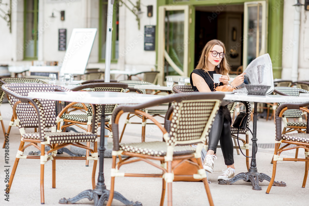 Young woman having a breakfast with coffee and croissant reading newspaper outdoors at the typical f